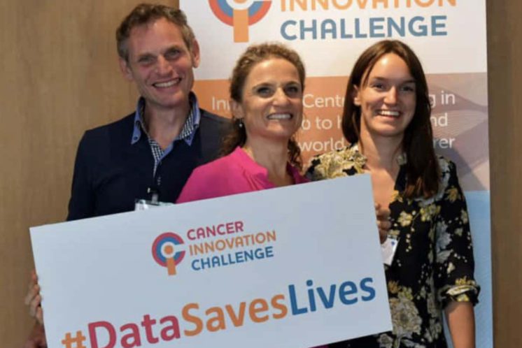 David and Anne holding a Cancer Innovation Challenge banner with #datasaveslives on the banner after winning prize money